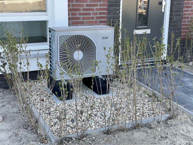 Beispiel einer Wärmepumpe Außeneinheit Wärmepumpe auf einem Kiesbett, umgeben von kleinen Pflanzen und einer Mauer.