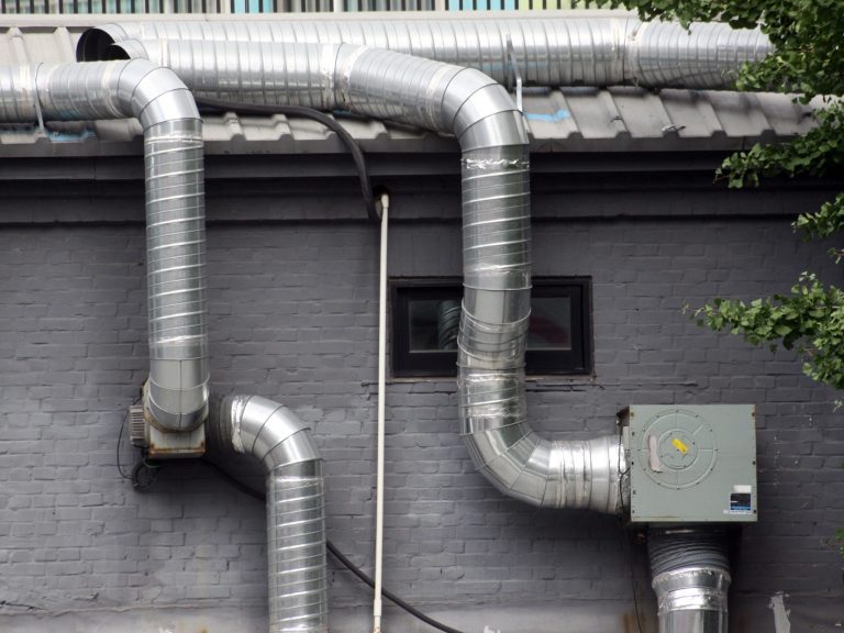 Lüftungsanlage Metallene Lüftungsrohre verlaufen an einer grauen Wand entlang, neben einem Fenster.